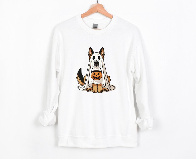 May include: White sweatshirt featuring a cartoon dog dressed as a ghost for Halloween. The dog is wearing a white sheet with eye holes and holding a jack-o'-lantern bucket. The dog has brown and black fur and is sitting down.