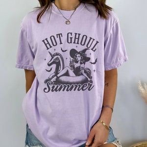 May include: A lavender t-shirt featuring a graphic of a witch riding a unicorn pool float. The text "Hot Ghoul Summer" is printed on the shirt.
