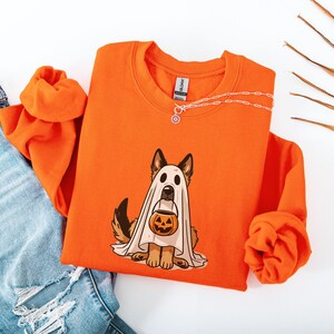 May include: Orange sweatshirt featuring a German Shepherd dressed as a ghost holding a jack-o'-lantern. The graphic is in the center of the shirt. A silver necklace is draped across the top of the shirt. Light blue jeans are visible in the image.