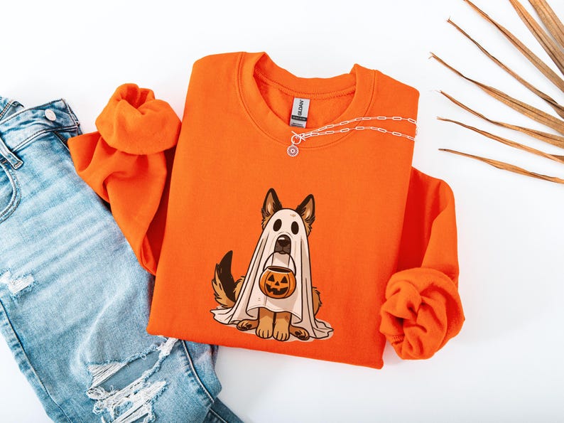 May include: Orange sweatshirt featuring a German Shepherd dressed as a ghost holding a jack-o'-lantern. The graphic is in the center of the shirt. A silver necklace is draped across the top of the shirt. Light blue jeans are visible in the image.
