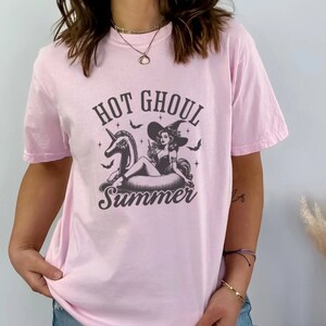 May include: Pink t-shirt featuring a black and white graphic of a witch riding a unicorn float with the text "Hot Ghoul Summer".