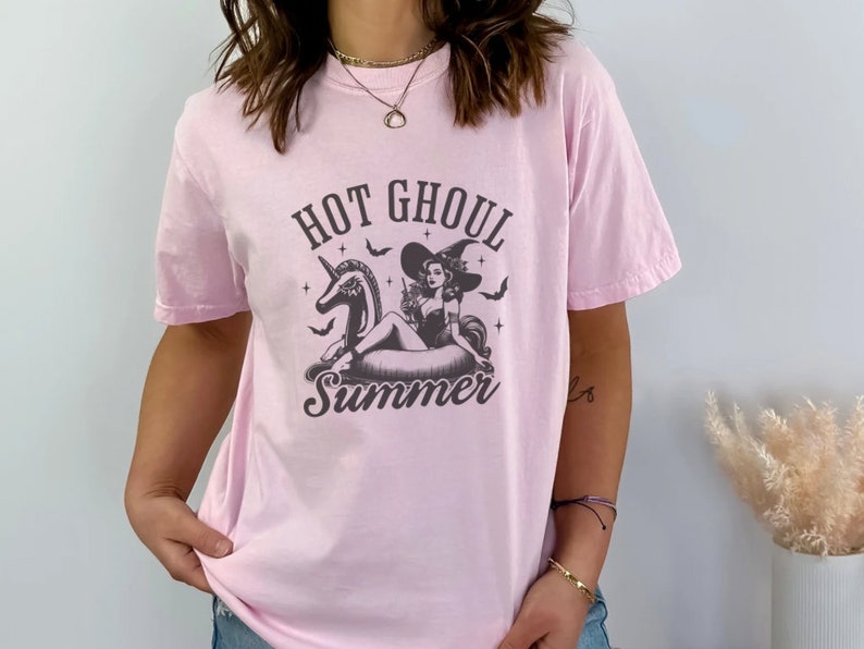 May include: Pink t-shirt featuring a black and white graphic of a witch riding a unicorn float with the text "Hot Ghoul Summer".