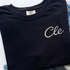 May include: Black Comfort Colors t-shirt with the word "Cle" embroidered in white pearls. The shirt is a crew neck and has a tag that says "Comfort Colors". The shirt is paired with blue denim jeans.