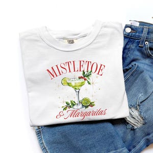 Pu&ograve; includere: Una t-shirt bianca con la grafica di un margarita verde in un bicchiere con fettine di lime, bacche rosse e foglie verdi. Il testo MISTLETOE & Margaritas &egrave; stampato in rosso sopra e sotto l'illustrazione del cocktail, circondato da stelle dorate.