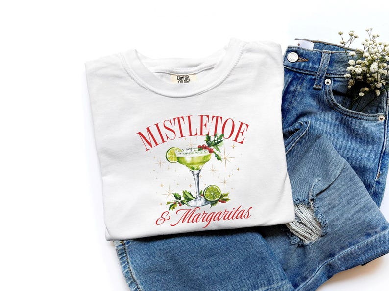 Pu&ograve; includere: Una t-shirt bianca con la grafica di un margarita verde in un bicchiere con fettine di lime, bacche rosse e foglie verdi. Il testo MISTLETOE & Margaritas &egrave; stampato in rosso sopra e sotto l'illustrazione del cocktail, circondato da stelle dorate.