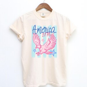 May include: Cream-colored t-shirt with a pink eagle graphic, blue stars, and the word "America" in blue script, along with the number "1776". The design is set against a light blue and white striped background.