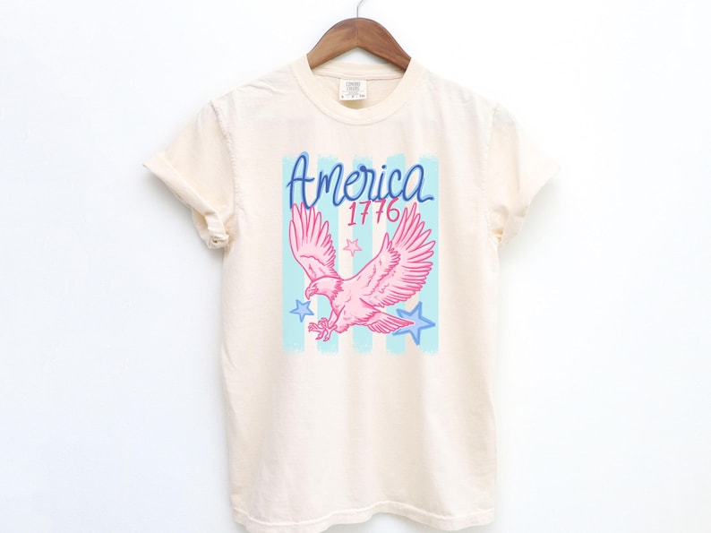 May include: Cream-colored t-shirt with a pink eagle graphic, blue stars, and the word "America" in blue script, along with the number "1776". The design is set against a light blue and white striped background.