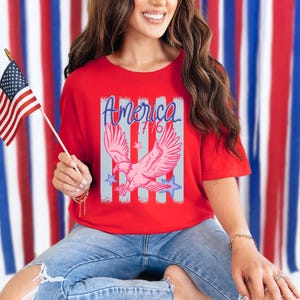 May include: Red t-shirt with a graphic of a pink eagle, the word "America" in blue script, and the number "76". The shirt is paired with blue jeans. An American flag is held in the hand.