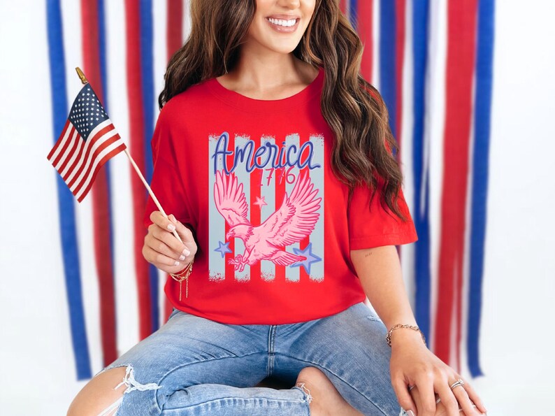 May include: Red t-shirt with a graphic of a pink eagle, the word "America" in blue script, and the number "76". The shirt is paired with blue jeans. An American flag is held in the hand.