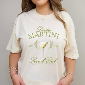 May include: A cream-coloured short-sleeve t-shirt featuring a graphic of a martini glass with olives. The text reads Dirty Martini Social Club, Extra Dirty, Extra Olives, and Vodka, Dry Vermouth, Olive Brine, Olives in a green serif font.