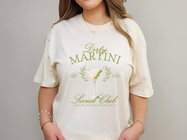 May include: A cream-coloured short-sleeve t-shirt featuring a graphic of a martini glass with olives. The text reads Dirty Martini Social Club, Extra Dirty, Extra Olives, and Vodka, Dry Vermouth, Olive Brine, Olives in a green serif font.