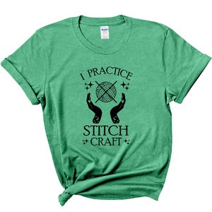 K&ouml;nnte beinhalten: Gr&uuml;nes T-Shirt mit dem Text "I practice stitch craft" in schwarzer Schrift. Der Text ist von Sternen und einer Grafik von H&auml;nden umgeben, die eine Wollkn&auml;uel halten.