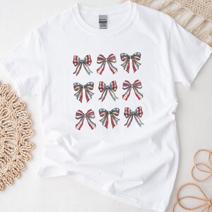 Puede incluir: Camiseta blanca con nueve dise&ntilde;os de lazos en rojo, blanco y azul, que recuerdan a la bandera estadounidense. Los lazos est&aacute;n dispuestos en un patr&oacute;n de cuadr&iacute;cula, a&ntilde;adiendo un toque patri&oacute;tico a la ropa casual.