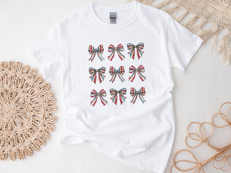 Puede incluir: Camiseta blanca con nueve dise&ntilde;os de lazos en rojo, blanco y azul, que recuerdan a la bandera estadounidense. Los lazos est&aacute;n dispuestos en un patr&oacute;n de cuadr&iacute;cula, a&ntilde;adiendo un toque patri&oacute;tico a la ropa casual.