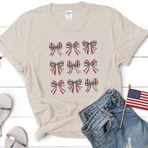 Puede incluir: Una camiseta color crema con nueve dise&ntilde;os de lazos en rojo, blanco y azul, que recuerdan la bandera estadounidense. La imagen incluye pantalones cortos de mezclilla, sandalias y gafas de sol rojas, lo que sugiere un tema patri&oacute;tico.