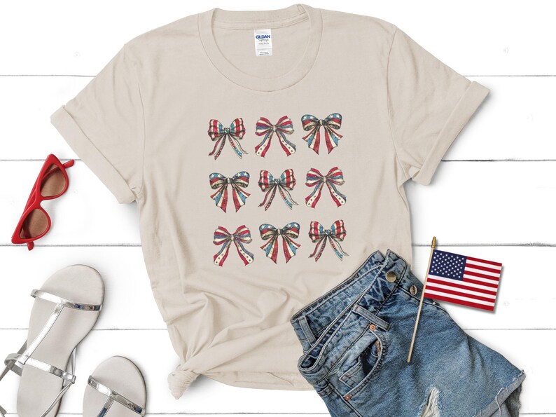 Puede incluir: Una camiseta color crema con nueve dise&ntilde;os de lazos en rojo, blanco y azul, que recuerdan la bandera estadounidense. La imagen incluye pantalones cortos de mezclilla, sandalias y gafas de sol rojas, lo que sugiere un tema patri&oacute;tico.