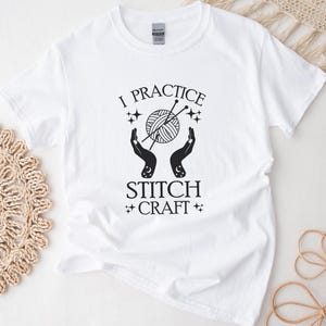 May include: White t-shirt with black text that reads "I practice stitch craft" with a graphic of two hands holding a ball of yarn and knitting needles.