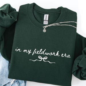 May include: A dark green sweatshirt with the text "in my fieldwork era" in white script. A silver necklace with a small pendant is draped across the front. The sweatshirt is folded with a pair of blue jeans.