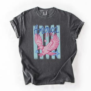 May include: Dark gray t-shirt featuring a pink eagle graphic with the word "America" and the number "1776" in blue. The design includes blue stars and vertical stripes, evoking a patriotic theme.