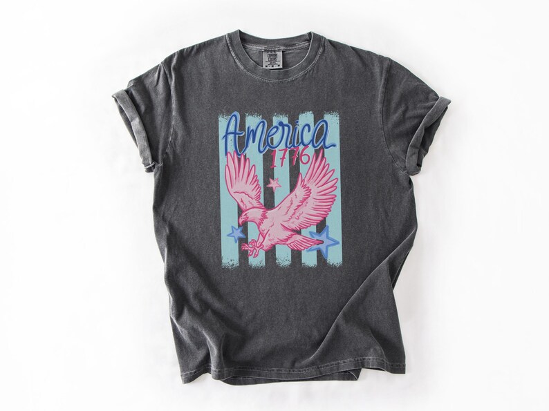 May include: Dark gray t-shirt featuring a pink eagle graphic with the word "America" and the number "1776" in blue. The design includes blue stars and vertical stripes, evoking a patriotic theme.