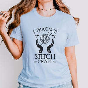 May include: A light blue t-shirt with the text "I practice stitch craft" in black. The design features a stylized image of a ball of yarn with knitting needles and two hands holding the yarn.