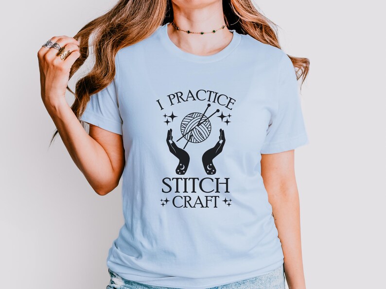 May include: A light blue t-shirt with the text "I practice stitch craft" in black. The design features a stylized image of a ball of yarn with knitting needles and two hands holding the yarn.
