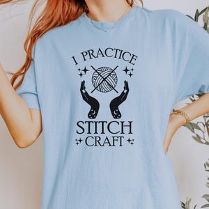 K&ouml;nnte beinhalten: Ein hellblaues T-Shirt mit einem schwarzen Grafikdesign, auf dem "I practice stitch craft" steht. Die Grafik zeigt eine Wollkn&auml;uel mit Stricknadeln und zwei H&auml;nde mit Sternen und Monden.