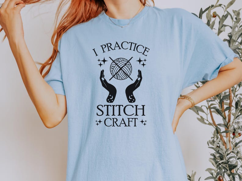 K&ouml;nnte beinhalten: Ein hellblaues T-Shirt mit einem schwarzen Grafikdesign, auf dem "I practice stitch craft" steht. Die Grafik zeigt eine Wollkn&auml;uel mit Stricknadeln und zwei H&auml;nde mit Sternen und Monden.