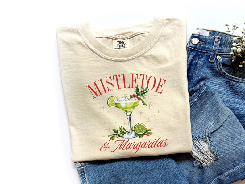 Pu&ograve; includere: Una t-shirt color crema con la grafica di un margarita verde in un bicchiere con una fetta di lime, bacche rosse e foglie verdi. Il testo MISTLETOE & MARGARITAS &egrave; stampato in rosso sopra e sotto l'illustrazione del cocktail.