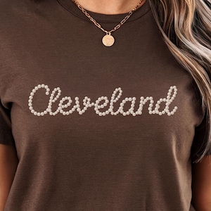 May include: A brown t-shirt with the name "Cleveland" spelled out in white, pearl-like beads. The shirt has short sleeves and a crew neck. A gold necklace with a pendant is visible.