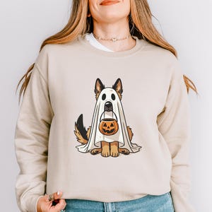 May include: Beige sweatshirt featuring a cartoon German Shepherd dressed as a ghost for Halloween. The dog is holding a jack-o'-lantern bucket. The design is centered on the front of the long-sleeved top.