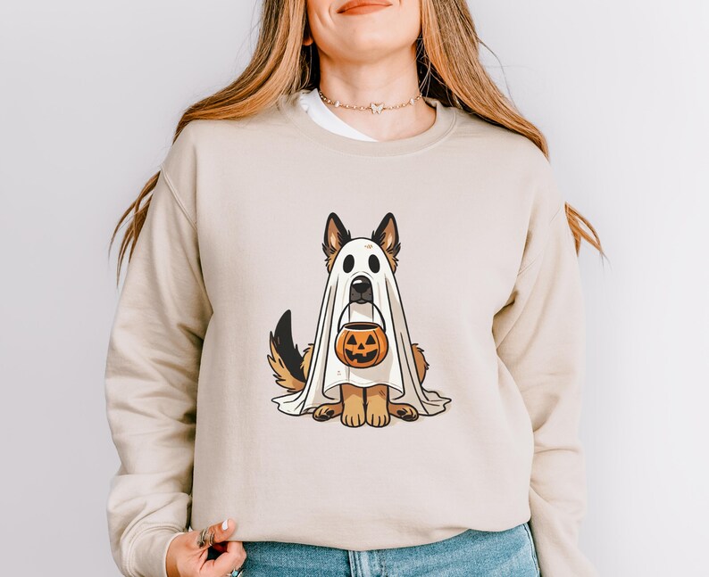May include: Beige sweatshirt featuring a cartoon German Shepherd dressed as a ghost for Halloween. The dog is holding a jack-o'-lantern bucket. The design is centered on the front of the long-sleeved top.