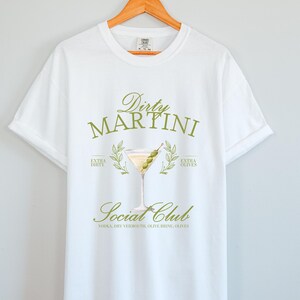 May include: A white short-sleeve t-shirt featuring a graphic of a martini glass with olives. The text reads Dirty Martini Social Club, Extra Dirty, Extra Olives, and Vodka, Dry Vermouth, Olive Brine, Olives in a muted olive green colour scheme.