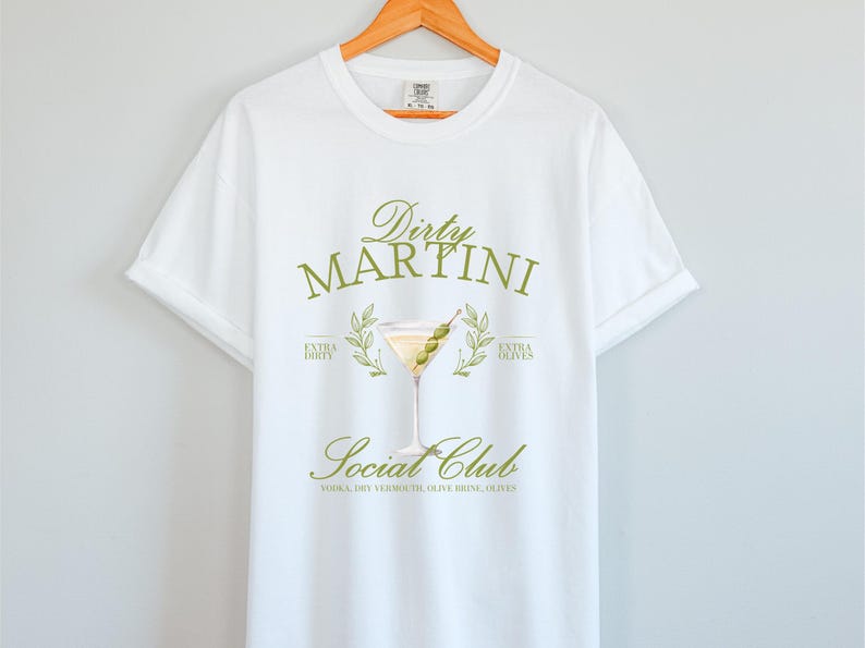 May include: A white short-sleeve t-shirt featuring a graphic of a martini glass with olives. The text reads Dirty Martini Social Club, Extra Dirty, Extra Olives, and Vodka, Dry Vermouth, Olive Brine, Olives in a muted olive green colour scheme.