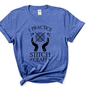 K&ouml;nnte beinhalten: Ein blaues T-Shirt mit dem Text "I practice stitch craft" in schwarzen Buchstaben. Der Text ist umgeben von einem stilisierten Bild eines Wollkn&auml;uels mit zwei H&auml;nden, die es halten.