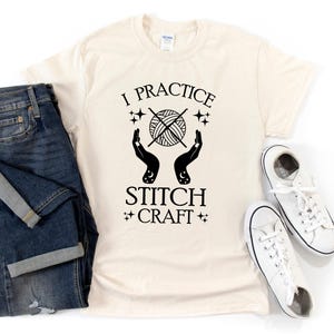 K&ouml;nnte beinhalten: Ein cremefarbenes T-Shirt mit schwarzem Text, der "I practice stitch craft" lautet. Der Text ist von Sternen und einer Grafik mit zwei H&auml;nden umgeben, die eine Wollkn&auml;uel mit Stricknadeln halten.