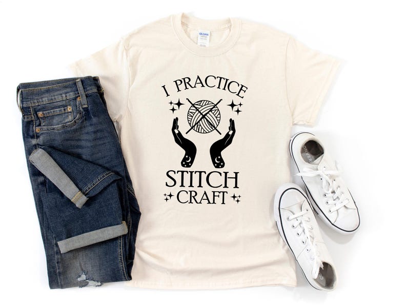 K&ouml;nnte beinhalten: Ein cremefarbenes T-Shirt mit schwarzem Text, der "I practice stitch craft" lautet. Der Text ist von Sternen und einer Grafik mit zwei H&auml;nden umgeben, die eine Wollkn&auml;uel mit Stricknadeln halten.