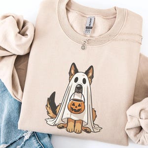 May include: Beige sweatshirt featuring a German Shepherd dog dressed as a ghost for Halloween. The dog is holding a jack-o'-lantern bucket. The sweatshirt has a crew neck and long sleeves. A silver necklace with a small pendant is draped on the shirt.