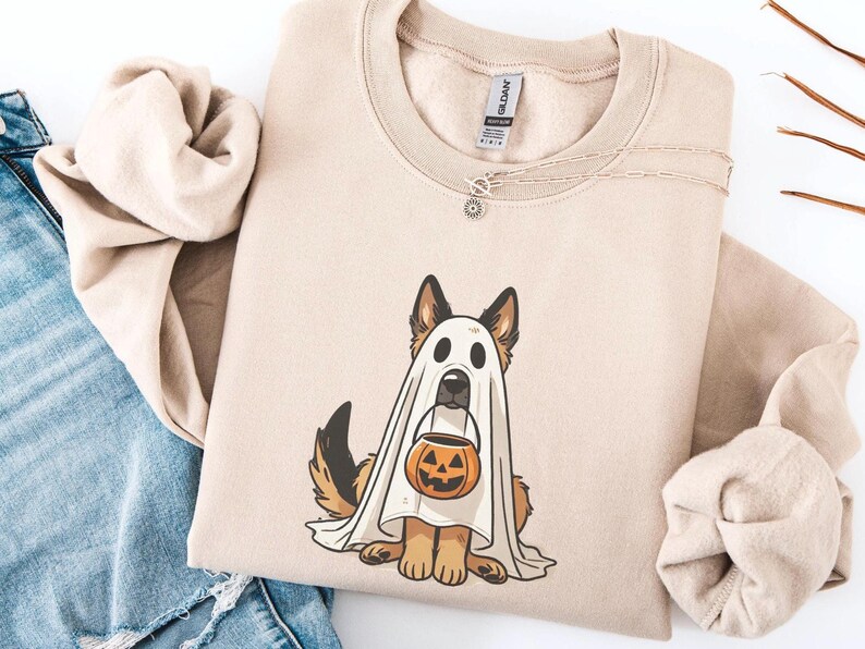 May include: Beige sweatshirt featuring a German Shepherd dog dressed as a ghost for Halloween. The dog is holding a jack-o'-lantern bucket. The sweatshirt has a crew neck and long sleeves. A silver necklace with a small pendant is draped on the shirt.