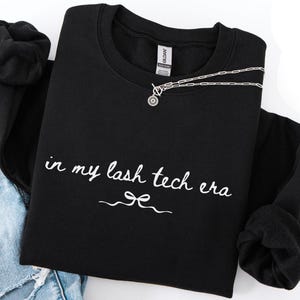 May include: Black crewneck sweatshirt with the text "in my lash tech era" in white script. A silver necklace with a circular pendant and a toggle clasp is draped across the sweatshirt. Rolled-up black sleeves and denim jeans are visible.