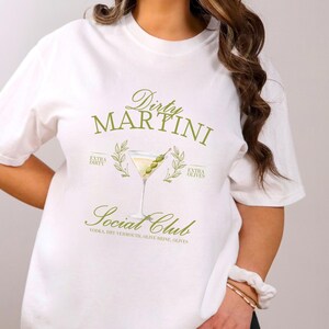 May include: A white short-sleeve t-shirt featuring a green graphic of a martini glass with olives. The text reads Dirty Martini Social Club, Extra Dirty, Extra Olives, and lists ingredients including vodka, dry vermouth, olive brine, and olives.