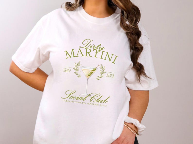 May include: A white short-sleeve t-shirt featuring a green graphic of a martini glass with olives. The text reads Dirty Martini Social Club, Extra Dirty, Extra Olives, and lists ingredients including vodka, dry vermouth, olive brine, and olives.