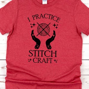 K&ouml;nnte beinhalten: Rotes T-Shirt mit schwarzem Text, der "I practice stitch craft" lautet. Der Text ist von einem stilisierten Bild eines Wollkn&auml;uels mit Stricknadeln und Sternen umgeben.