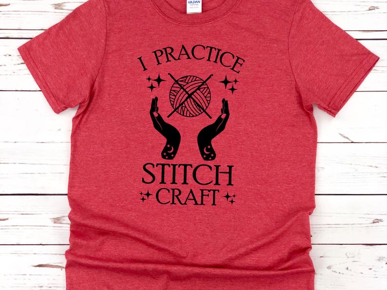 K&ouml;nnte beinhalten: Rotes T-Shirt mit schwarzem Text, der "I practice stitch craft" lautet. Der Text ist von einem stilisierten Bild eines Wollkn&auml;uels mit Stricknadeln und Sternen umgeben.