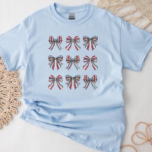 Puede incluir: Camiseta azul claro con nueve dise&ntilde;os de lazos en rojo, blanco y azul, que recuerdan a la bandera estadounidense. Los lazos tienen un efecto acuarela, a&ntilde;adiendo un toque suave al tema patri&oacute;tico. La camiseta es de material suave.