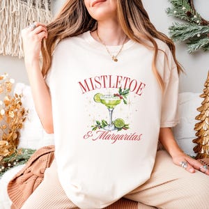 Pu&ograve; includere: Una t-shirt color crema con la grafica di un bicchiere da margarita guarnito con una fetta di lime e vischio. Il testo sopra il bicchiere recita MISTLETOE e il testo sottostante recita & Margaritas in un carattere rosso con grazie.