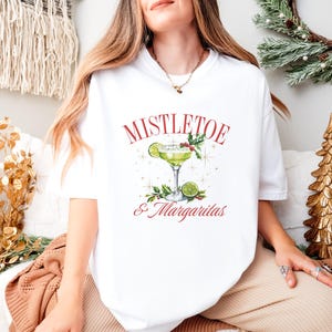 Pu&ograve; includere: Una t-shirt bianca a maniche corte con la grafica di un margarita verde in un bicchiere con fettine di lime, foglie di agrifoglio e bacche. Il testo MISTLETOE & MARGARITAS &egrave; stampato in rosso con piccoli accenti a forma di stella dorata.