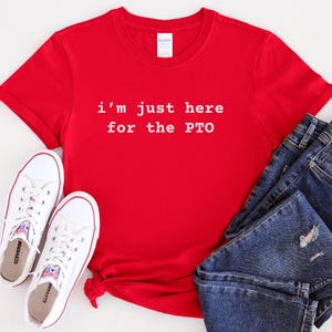 May include: Red t-shirt with white text that reads "i'm just here for the PTO". The shirt is laid flat on a white surface with a pair of white sneakers and blue jeans.