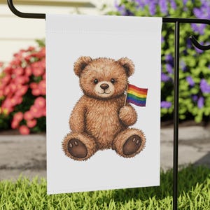 Puede incluir: Una bandera de jardín blanca con un oso de peluche marrón sosteniendo una bandera arcoíris. El oso tiene una expresión amigable. La bandera se muestra en un soporte de metal negro, sobre un fondo de flores coloridas y hierba verde.