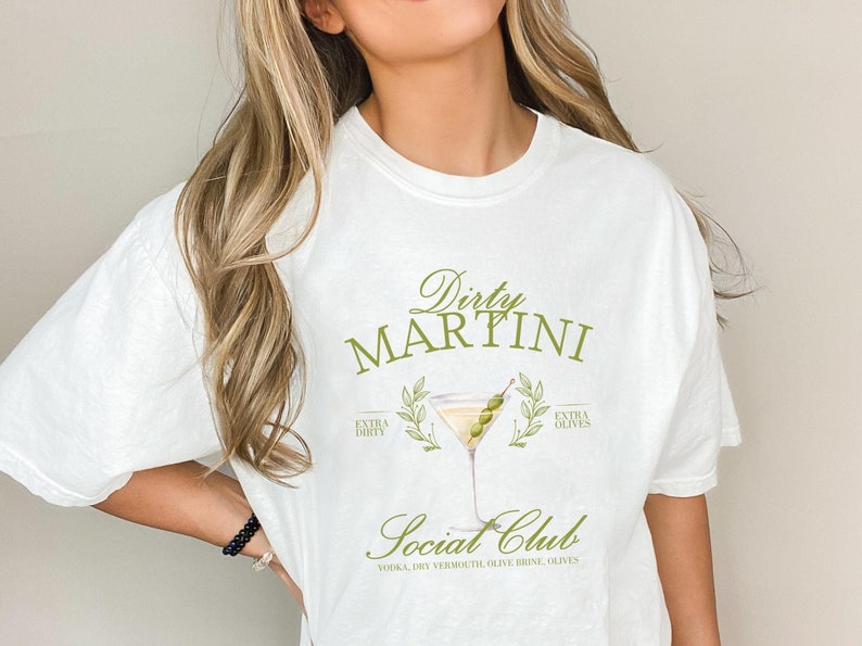 May include: A white t-shirt featuring a green graphic print of a martini glass with three olives. The text reads Dirty MARTINI Social Club, Extra Dirty, Extra Olives, and Vodka, Dry Vermouth, Olive Brine, Olives in a decorative green font.